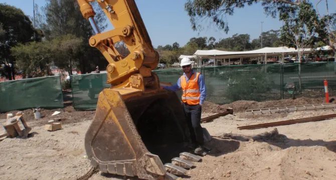 An excavator bucket resting on a construction site with a UDEC worker in hi-vis and hard hat alongside